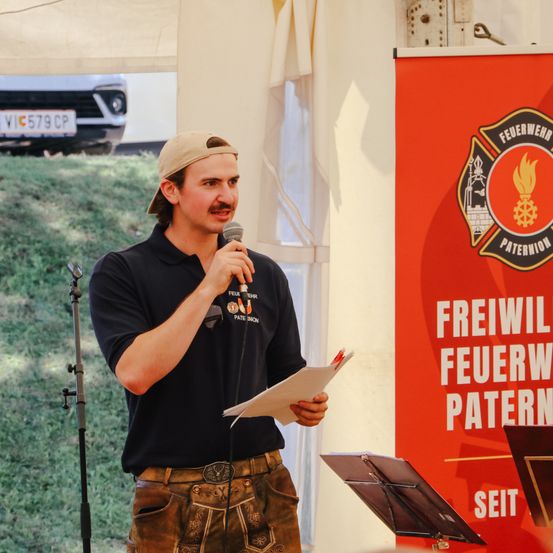 Ein Mann in einer Mütze und einem Polohemd spricht in ein Mikrofon, während er ein Papier und einen Mikrofonständer in einem Zelt hält.