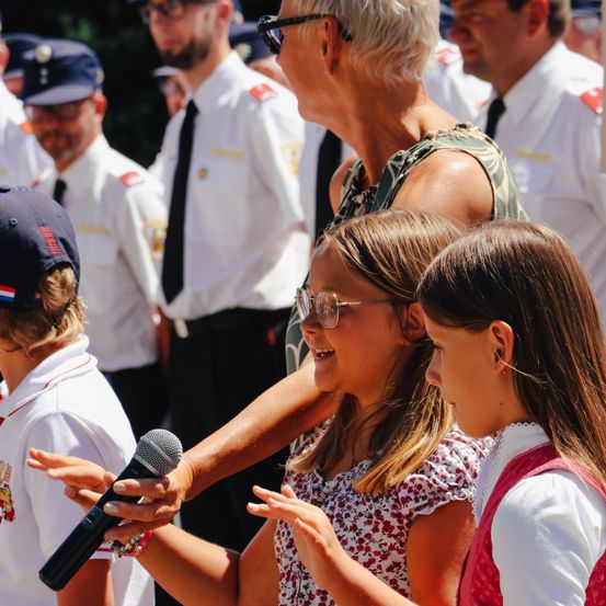 Eine Frau hält ein Mikrofon, während sie ein junges Mädchen führt, das hineinspricht. Sie stehen neben einem Mädchen mit langen Haaren. Hinter ihnen sind Menschen in Uniformen.