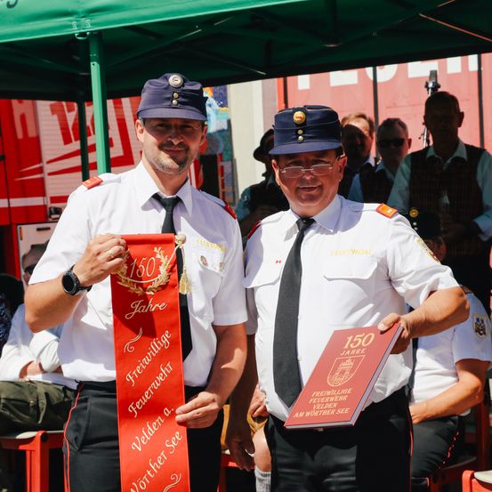 Zwei uniformierte Männer stehen unter einem Zelt, lächelnd und halten eine rote Schleife und ein Buch, und feiern 150 Jahre freiwillige Feuerwehrleute in Wörther See.