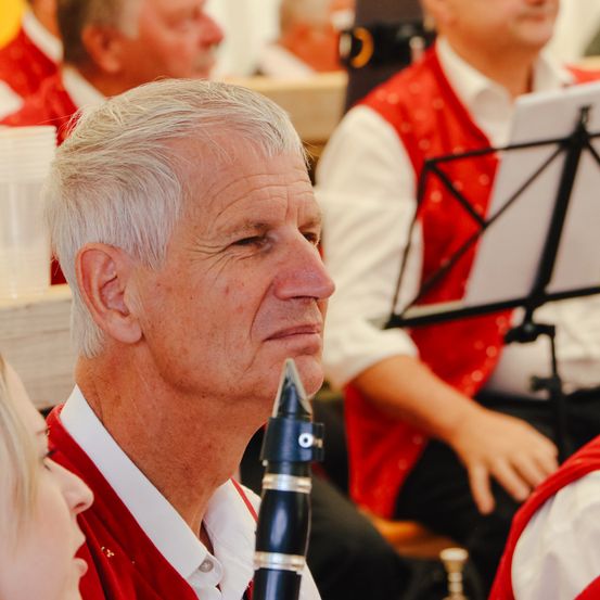 Ein älterer Mann mit grauem Haar spielt Klarinette. Er trägt eine rote Weste und ein weißes Hemd. Hinter ihm ist ein weiterer Mann, der ebenfalls eine rote Weste trägt. Sie sind wahrscheinlich bei einer Bandprobe.