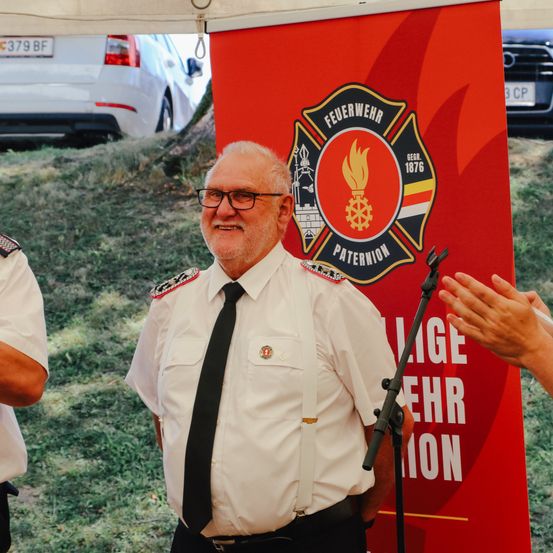 Ein lächelnder Mann in einer Feuerwehruniform steht vor einem Banner, während eine andere Person klatscht. Ein Auto ist hinter dem Banner geparkt.
