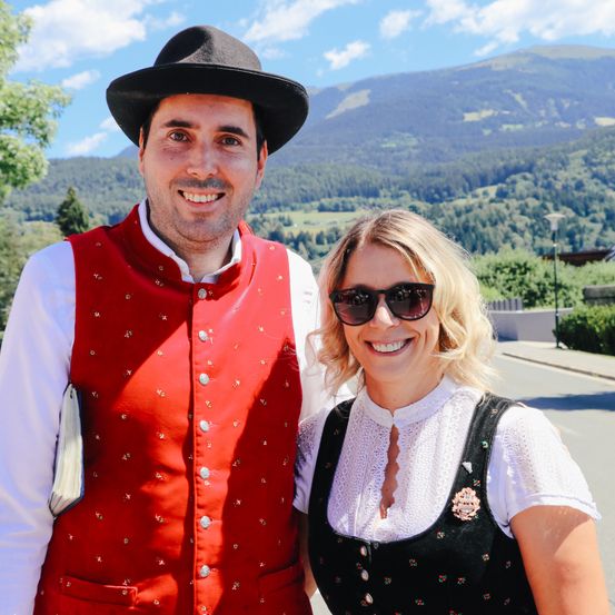 Ein lächelndes Paar steht vor einer Bergkulisse in traditioneller Kleidung, der Mann in einer roten Weste und Hut und die Frau in einem weißen Blusenkleid mit schwarzer Schürze.
