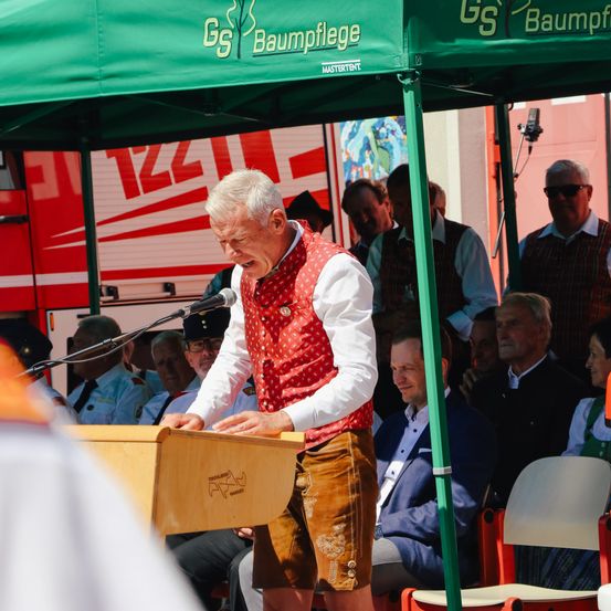 Ein älterer Mann mit einem roten Westchen steht hinter einem Pult unter einem grünen Zelt und spricht in ein Mikrofon. Hinter ihm sitzen und stehen mehrere Menschen, darunter ein Mann in einem weißen Hemd.
