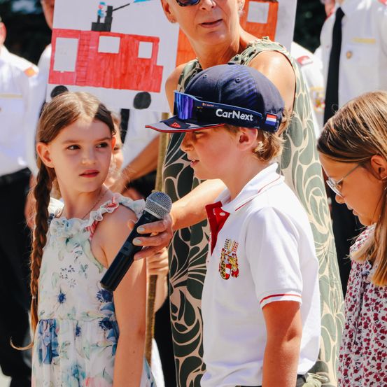 Ein Junge mit einer CarNext-Kappe hält ein Mikrofon an ein Mädchen in einem Blumenkleid, eine erwachsene Frau hält ein Schild hinter ihnen.
