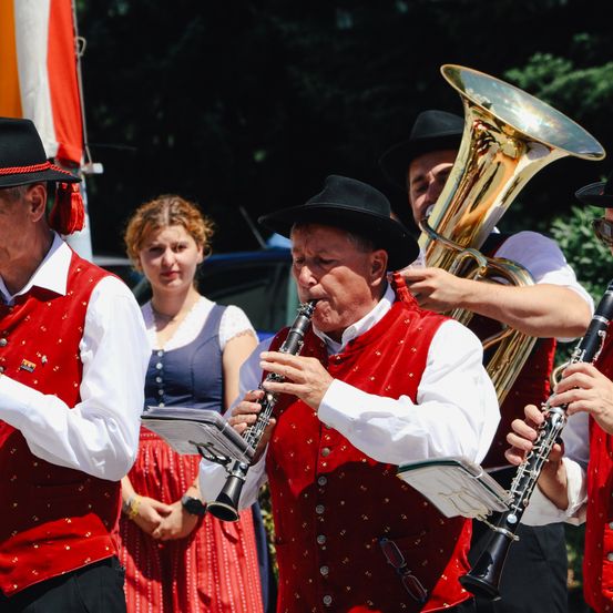 Eine Gruppe Musiker in traditioneller Kleidung spielt Instrumente im Freien, mit einer Frau und Bäumen im Hintergrund.