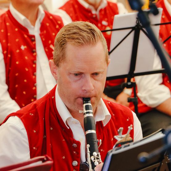 Ein Mann in einer roten Weste spielt Klarinette vor einem Notenständer, mit anderen Musikern im Hintergrund.