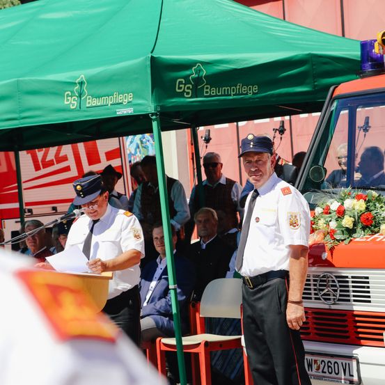 Ein Feuerwehrmann steht neben einem Feuerwehrauto unter einem grünen Zelt mit dem GS Baumpflege-Logo. Ein Mann liest eine Rede in ein Mikrofon. Leute sitzen im Hintergrund, manche tragen Hüte.