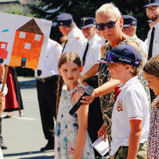 Ein junges Mädchen hält ein Mikrofon und ein Schild mit einem Haus-Zeichnung neben einem Jungen, der eine CarNext-Kappe trägt. Dahinter stehen mehrere uniformierte Erwachsene und beobachten.