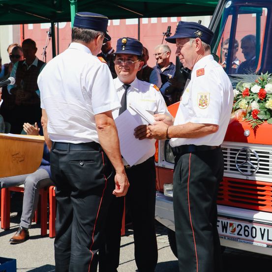 Drei Männer in Uniformen stehen vor einem Feuerwehrauto, einer hält ein Papier und spricht. Ein Publikum sitzt hinter ihnen unter einem Zelt.