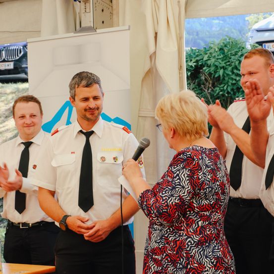 Eine Frau spricht in ein Mikrofon bei einer Veranstaltung. Ein Mann in Uniform steht neben ihr, und zwei weitere Männer stehen hinter ihm. Sie klatschen.