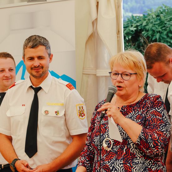 Eine Frau spricht in ein Mikrofon, während ein Feuerwehrmann hinter ihr steht. Dahinter ist ein weißer Hintergrund mit einem Logo. Ein Mann schaut lächelnd zu.