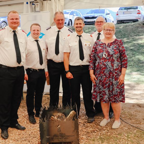 Fünf Personen in Uniformen stehen draußen in der Nähe eines Zeltes. Sie posieren für ein Foto mit einem metallischen Feuerkorb in der Mitte. Hinter ihnen sind Autos auf einer grasbewachsenen Anhöhe geparkt.