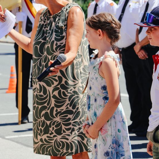 Eine erwachsene Frau spricht in ein Mikrofon, während ein junges Mädchen zuhört. Sie sind von anderen in Uniform umgeben. Ein Verkehrskegel ist im Hintergrund sichtbar.