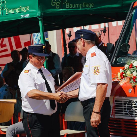 Zwei Feuerwehrleute in Uniform schütteln sich die Hände vor einer Menschenmenge und einem geschmückten Feuerwehrwagen.