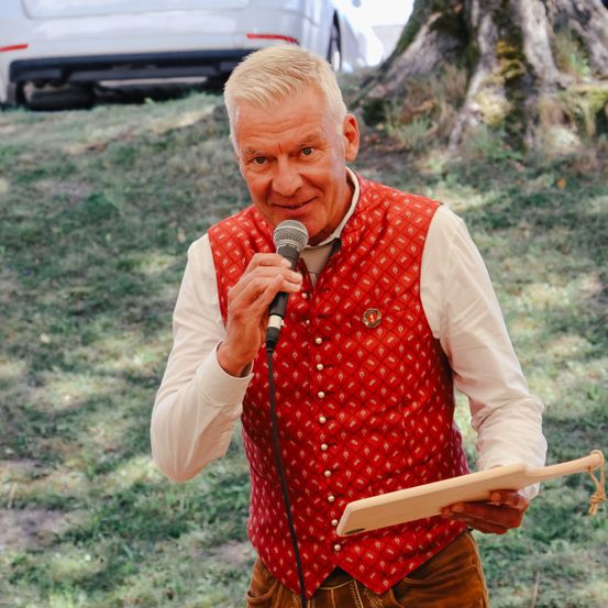 Ein Mann in einem roten Westchen und brauner Hose hält ein Mikrofon und eine Holztafel in einer grasbewachsenen Gegend. Ein Auto ist im Hintergrund geparkt.