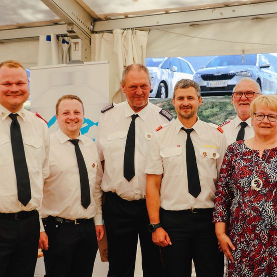 Fünf Menschen in Uniform und eine Frau stehen zusammen in einem Zelt und lächeln für ein Foto. Hinter ihnen ist ein Bild eines Autos.