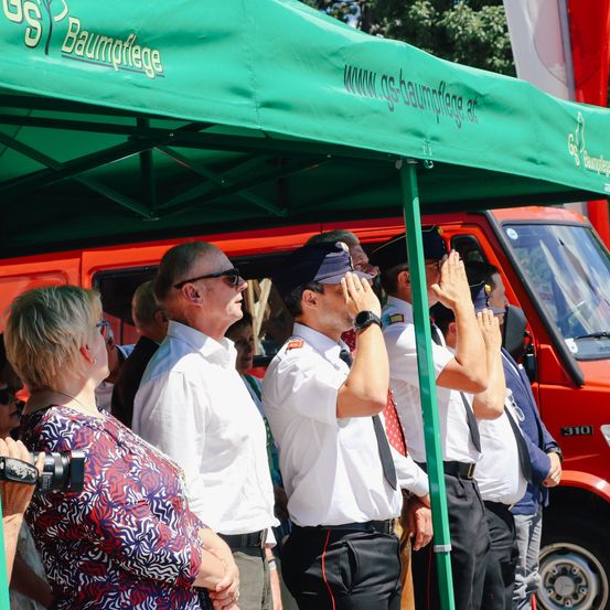 Mehrere Männer in Uniform salutieren unter einem grünen Zelt. Eine Frau beobachtet sie. Ein roter Van steht hinter ihnen.