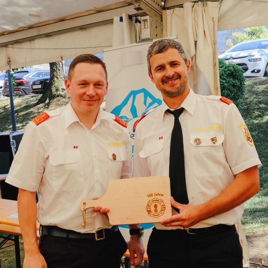 Zwei Männer in Uniformen posieren für ein Foto mit einer Holzplakette. Sie stehen draußen unter einem Zelt, mit einem Banner und geparkten Autos im Hintergrund.