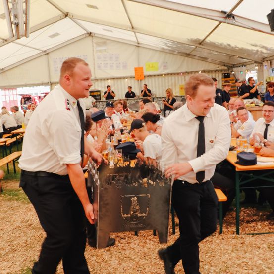 Zwei Männer in weißen Uniformen und Krawatten halten einen Grill mit Flammen in der Hand und gehen in einem Zelt im Freien. Viele Menschen sitzen um Tische, einige in Hüten. Ein Lautsprecher ist im Hintergrund.