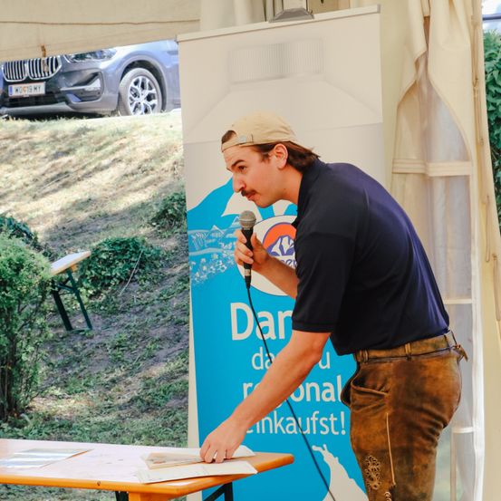 Ein Mann mit braunen Hosen und einer Mütze spricht in ein Mikrofon bei einer Veranstaltung im Freien, mit einem Auto und einem Schild im Hintergrund.