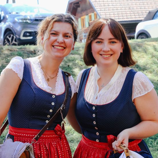 Zwei Frauen in traditionellen blauen gepunkteten Kleidern posieren für ein Foto im Freien mit Autos und einem Haus im Hintergrund.