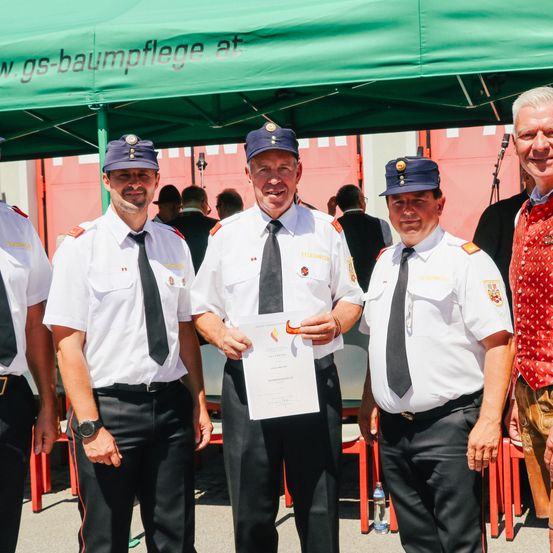 Fünf Männer in Uniform stehen unter einem grünen Zelt. Der Mann in der Mitte hält ein Zertifikat. Die Männer lächeln und scheinen für ein Foto zu posieren.