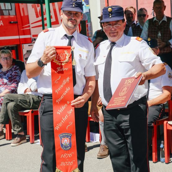 Zwei Männer in Uniformen halten Banner in der Hand und stehen vor einer Menge. Einer hält ein rotes Banner mit den Jahren 1876 und 2026. Der andere hält ein Zertifikat. Hinter ihnen sitzen Menschen auf Stühlen, und ein roter Feuerwehrwagen ist geparkt.