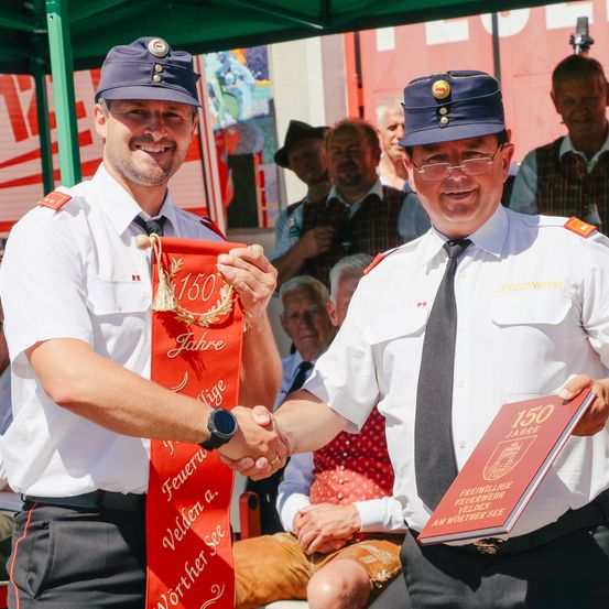 Zwei Männer in Uniformen schütteln sich die Hände, einer hält eine rote Schärpe mit '150 Jahre' und der andere hält ein rotes Buch. Sie stehen vor einer Menschenmenge.