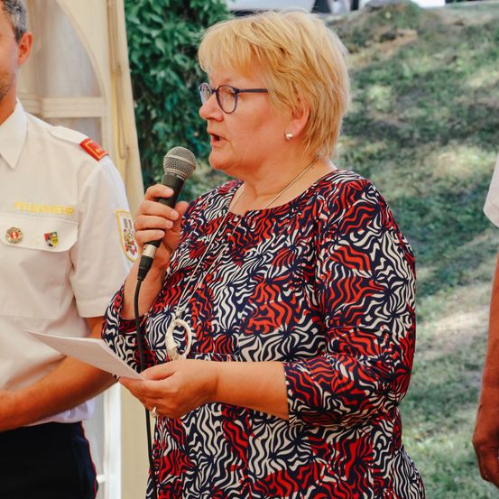 Eine Frau mit Brille spricht in ein Mikrofon, hält ein Blatt Papier in der Hand, mit einem Feuerwehrmann hinter ihr.