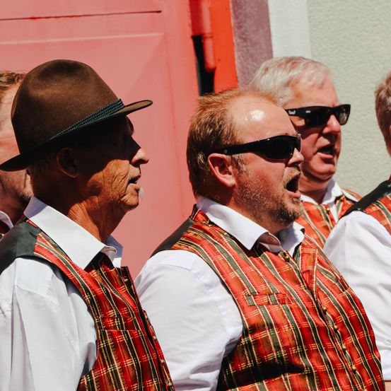 Vier ältere Männer, die karierte Westen und Hüte tragen, stehen zusammen und singen. Einer trägt eine Sonnenbrille. Sie stehen vor einer roten Wand.