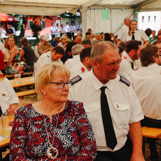 Eine Gruppe von Menschen in Uniformen sitzt in einem Zelt. Ein Mann und eine Frau sitzen eng beieinander und blicken in die gleiche Richtung. Es gibt Tische mit Gläsern und Stühlen.