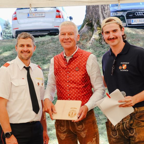 Drei Männer stehen im Freien, zwei in Uniformen. Der Mann in der Mitte hält eine Urkunde, und der Mann auf der linken Seite lächelt. Hinter ihnen sind Autos und Bäume zu sehen.