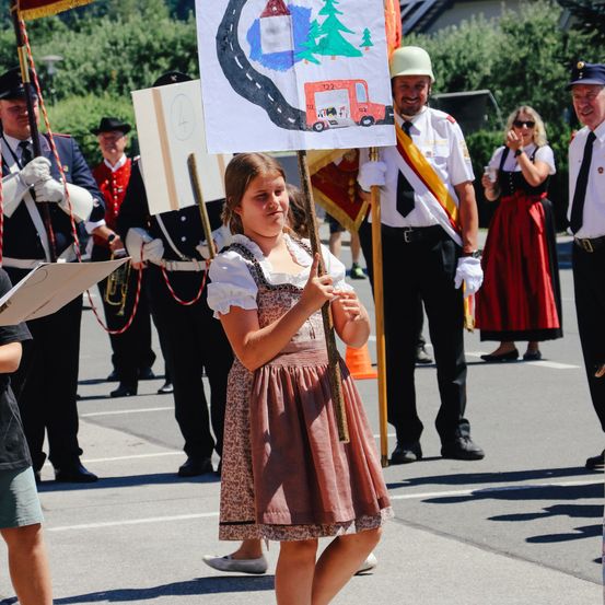 Ein Mädchen in einer traditionellen Kleidung hält ein Banner mit einer Straßenzeichnung und steht in einem Umzug mit Mitgliedern einer Blaskapelle. Dahinter gehen andere in Uniformen und traditioneller Kleidung in einem Umzug.