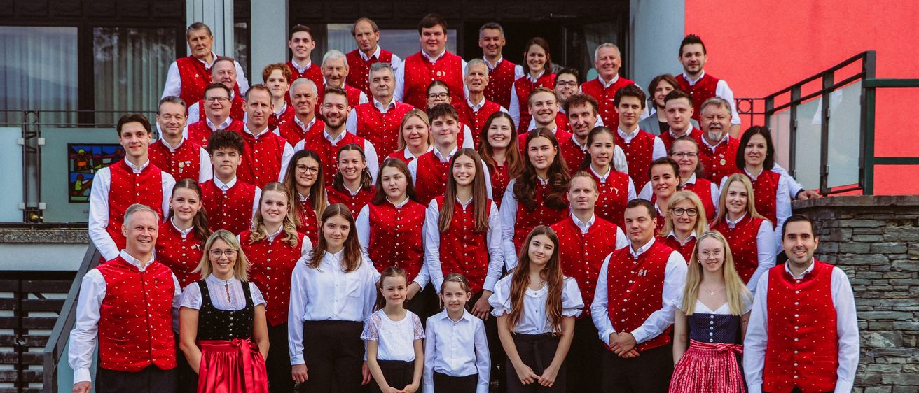 Eine große Gruppe von Menschen in roten Westen und weißen Hemden steht auf einer Treppe vor einem Gebäude. Einige sind Kinder, und sie alle lächeln.