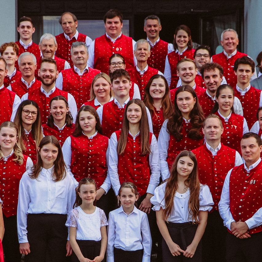 Eine große Gruppe von Menschen in roten Westen und weißen Hemden steht auf einer Treppe vor einem Gebäude. Einige sind Kinder, und sie alle lächeln.