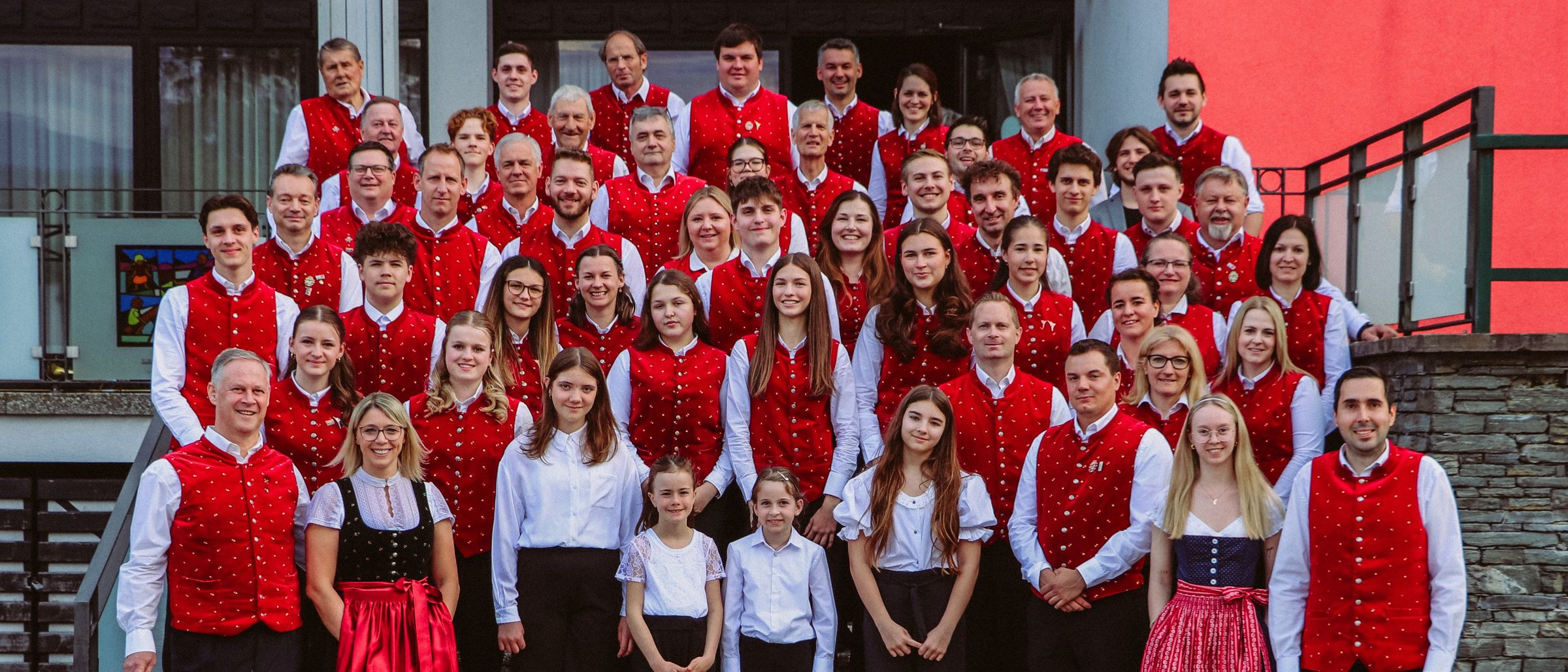 Eine große Gruppe von Menschen in roten Westen und weißen Hemden steht auf einer Treppe vor einem Gebäude. Einige sind Kinder, und sie alle lächeln.