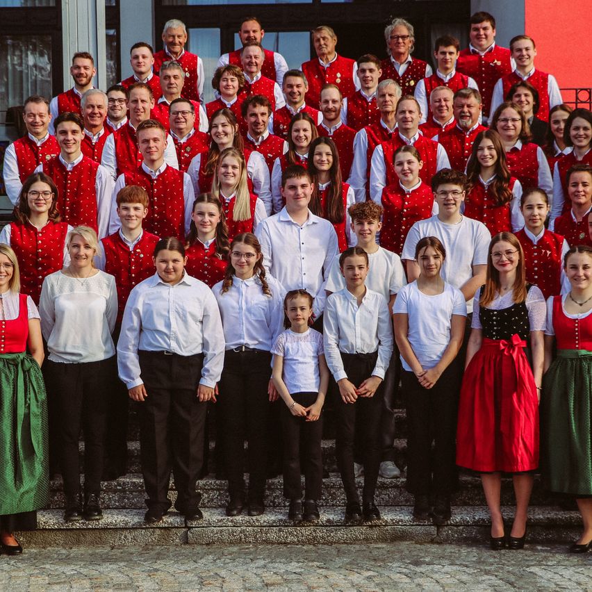 Ein Chor steht auf den Stufen eines Gebäudes und trägt rote Westen mit weißen Besätzen. Einige von ihnen tragen Brillen. Das Gebäude hat eine rote Wand.