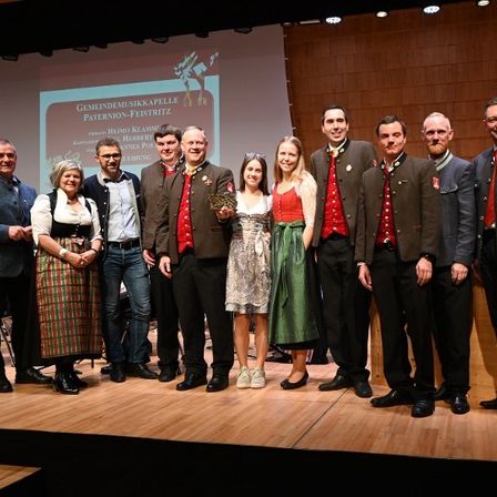 Gruppe von Erwachsenen, meist in traditioneller Kleidung, steht für ein Foto auf der Bühne. Eine Leinwand im Hintergrund zeigt Text zu einer Musikveranstaltung.