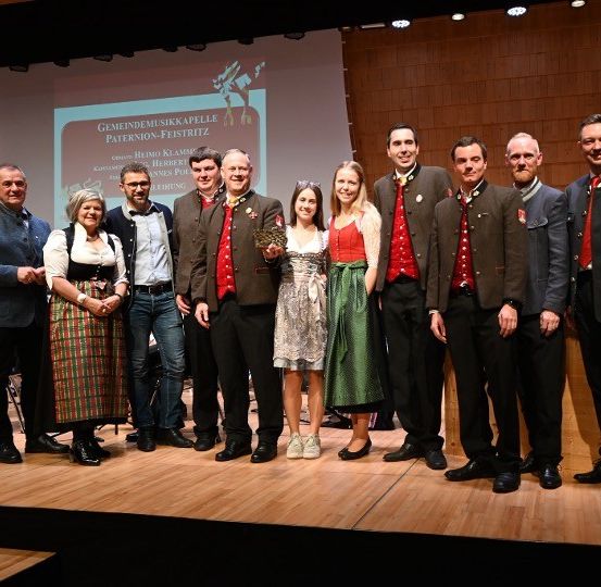 Gruppe von Erwachsenen, meist in traditioneller Kleidung, steht für ein Foto auf der Bühne. Eine Leinwand im Hintergrund zeigt Text zu einer Musikveranstaltung.