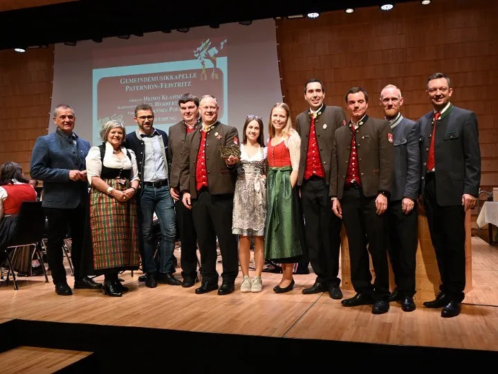 Gruppe von Erwachsenen, meist in traditioneller Kleidung, steht für ein Foto auf der Bühne. Eine Leinwand im Hintergrund zeigt Text zu einer Musikveranstaltung.