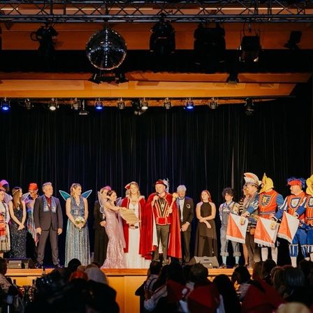 Eine Gruppe von Menschen in verschiedenen Kostümen steht auf der Bühne, wahrscheinlich bei einer Aufführung oder einem Fest. Einige halten Fahnen, und eine Person sticht in einem roten Umhang hervor. Scheinwerfer und eine Discokugel hängen von der Decke.