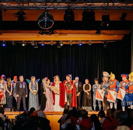 Eine Gruppe von Menschen in verschiedenen Kostümen steht auf der Bühne, wahrscheinlich bei einer Aufführung oder einem Fest. Einige halten Fahnen, und eine Person sticht in einem roten Umhang hervor. Scheinwerfer und eine Discokugel hängen von der Decke.