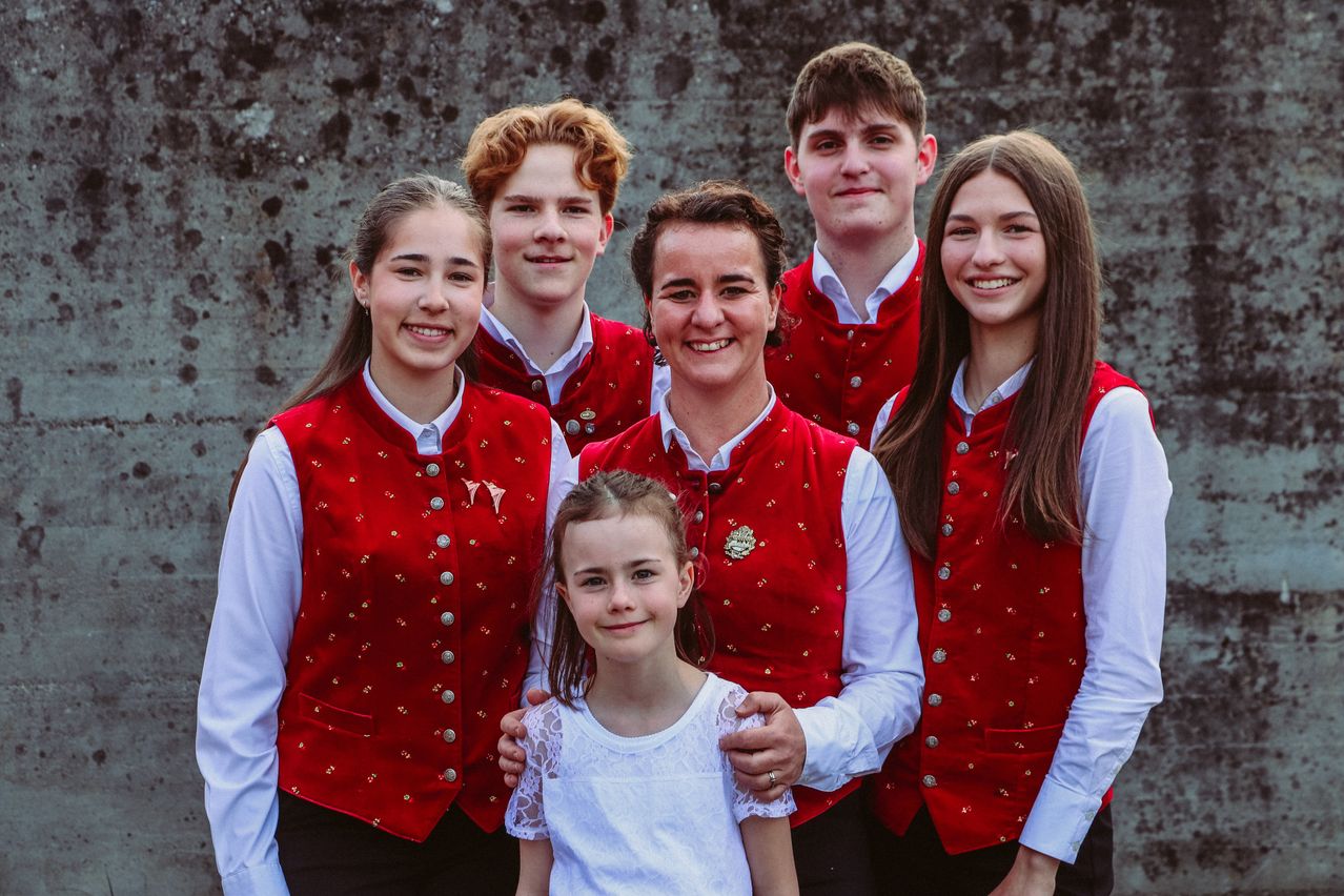 Eine Familiengruppe steht gemeinsam in traditionellen roten Westen und lächelt für ein Foto. Die Frau in der Mitte hält ein junges Mädchen auf dem Schoß. Dahinter befindet sich eine strukturierte Wand.