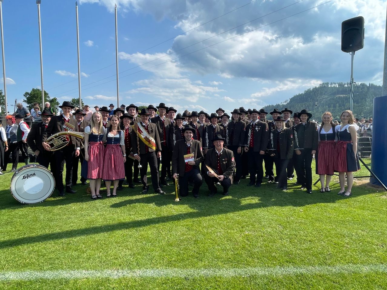 Eine große Gruppe von Menschen in traditioneller Kleidung und mit Musikinstrumenten posiert für ein Foto auf einem Rasenfeld mit Bergen im Hintergrund.