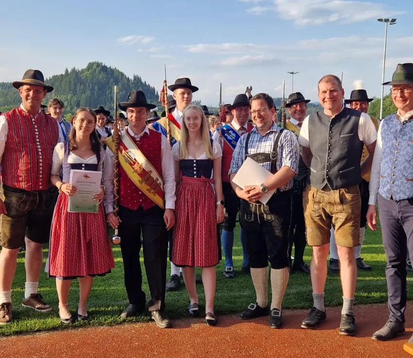 Eine Gruppe von Menschen in traditioneller bayerischer Kleidung posiert für ein Foto auf einer Rasenfläche mit Bergen im Hintergrund. Zwei Frauen halten Urkunden, und ein Mann hält Papiere.