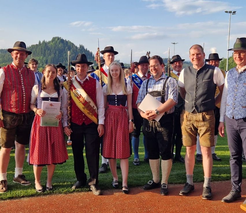 Eine Gruppe von Menschen in traditioneller bayerischer Kleidung posiert für ein Foto auf einer Rasenfläche mit Bergen im Hintergrund. Zwei Frauen halten Urkunden, und ein Mann hält Papiere.