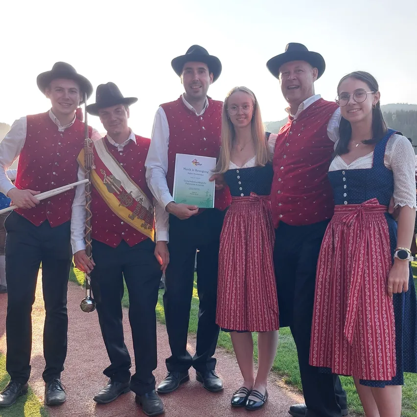 Eine Gruppe von Personen in traditioneller Kleidung posiert für ein Foto. Ein Mann in der Mitte hält ein Zertifikat. Im Hintergrund befindet sich eine Rasenfläche und in der Ferne Berge.