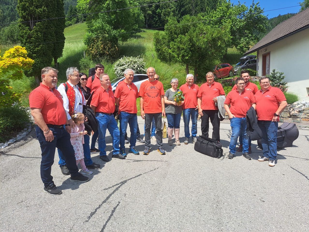 Eine Gruppe von Erwachsenen und Kindern in roten Shirts, versammelt in einer malerischen Außenumgebung mit Bäumen und einem Haus. Sie lächeln und scheinen für ein Foto zu posieren.