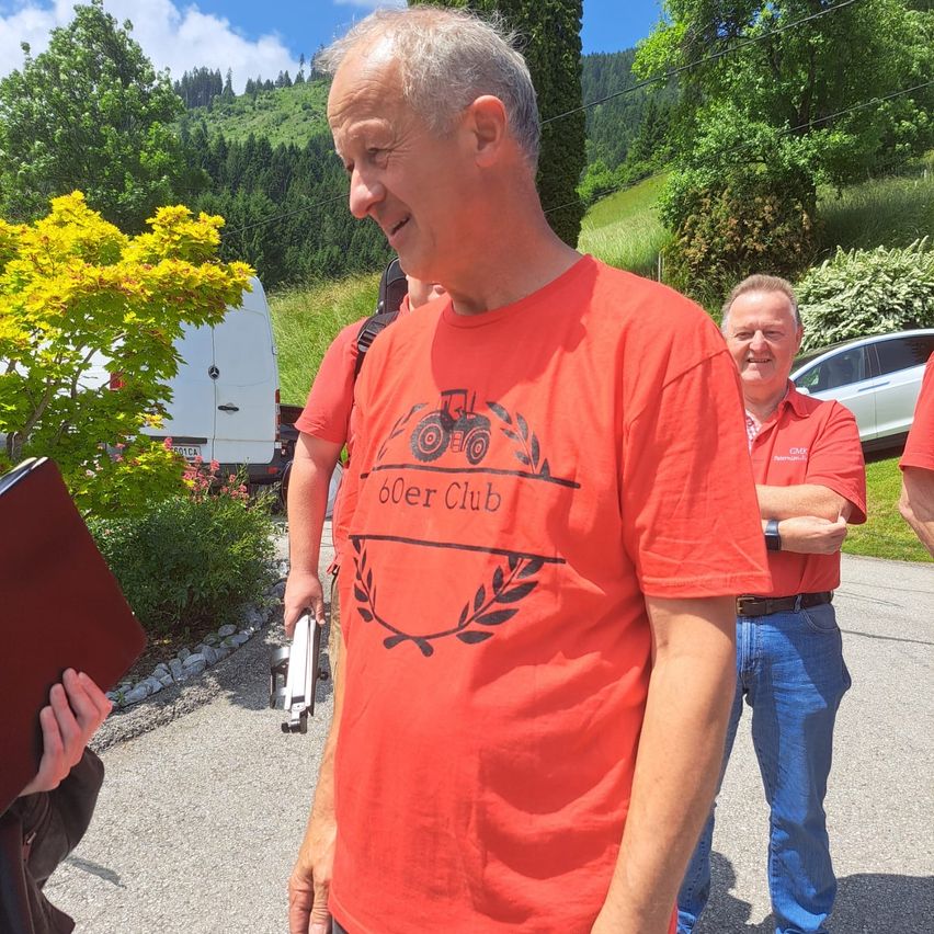 Ein Mann in einem roten T-Shirt steht lächelnd, mit zwei anderen hinter ihm, einer hält eine Mappe. Sie sind in einer malerischen Außenumgebung mit einem Berg, Bäumen und geparkten Fahrzeugen.