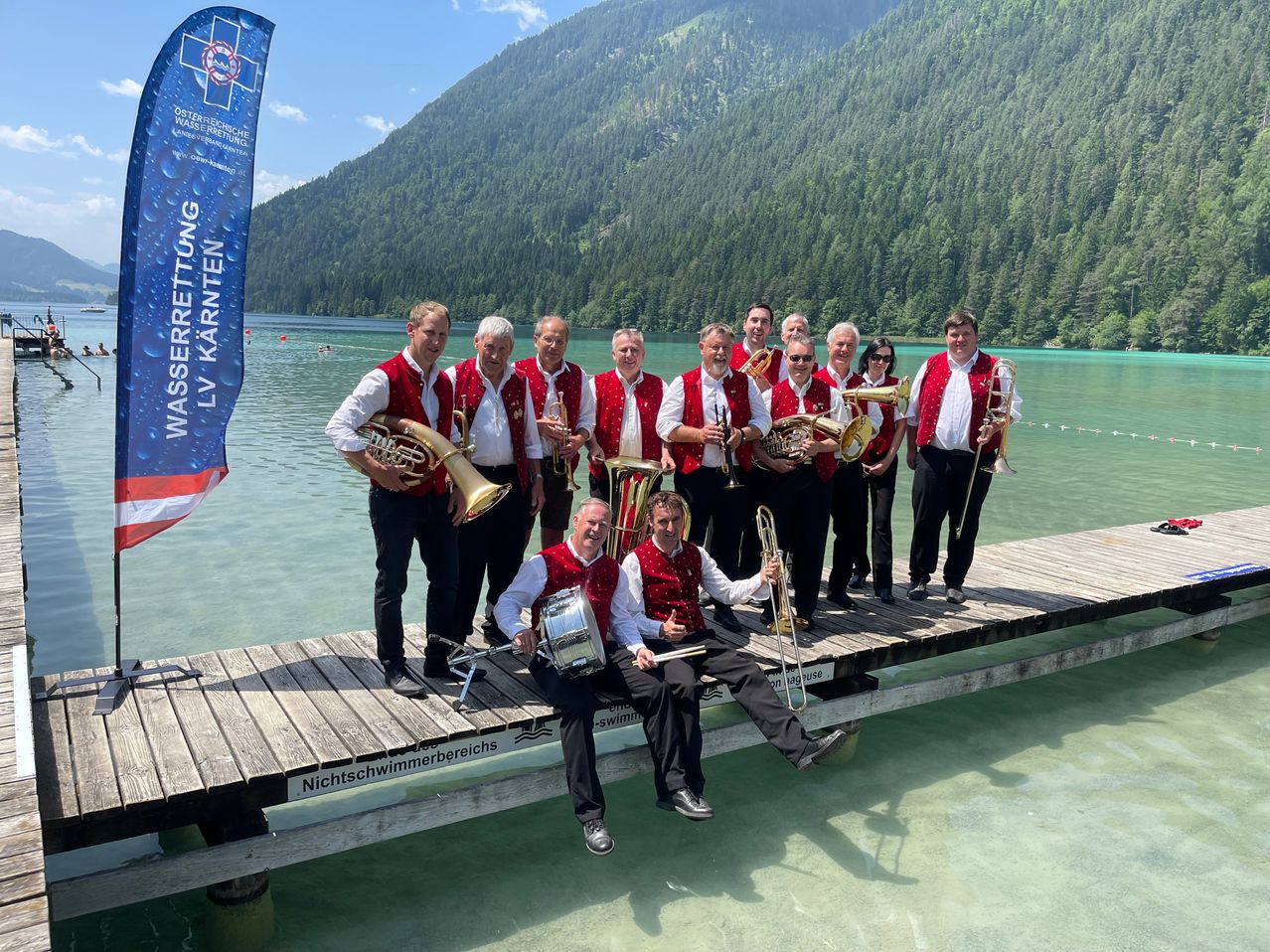 Eine Brassband posiert auf einer Pier am See, mit Bergen im Hintergrund. Sie tragen rote Westen und halten verschiedene Instrumente.